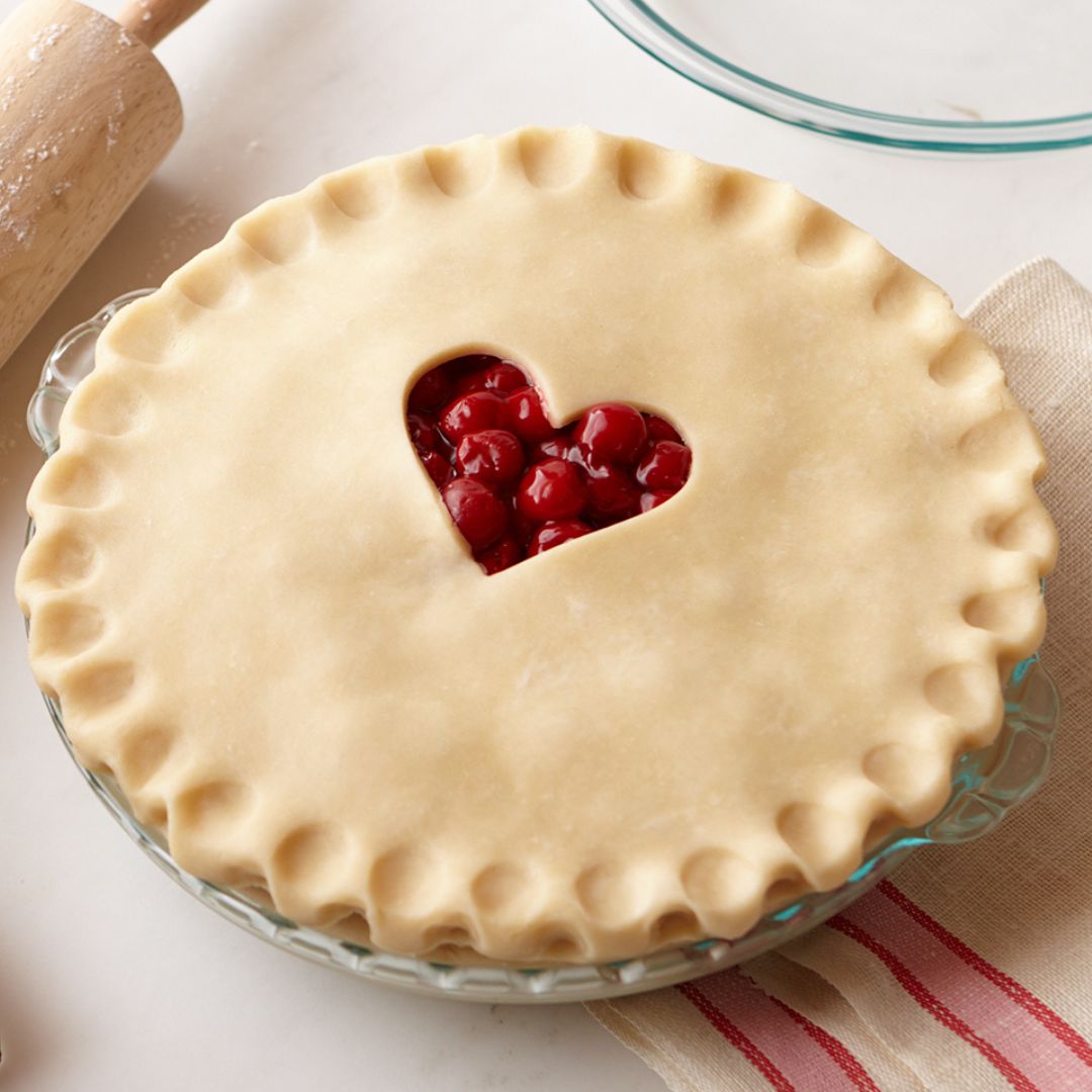 Pie with a heart-shaped cutout on top, filled with red fruit, on a white surface.