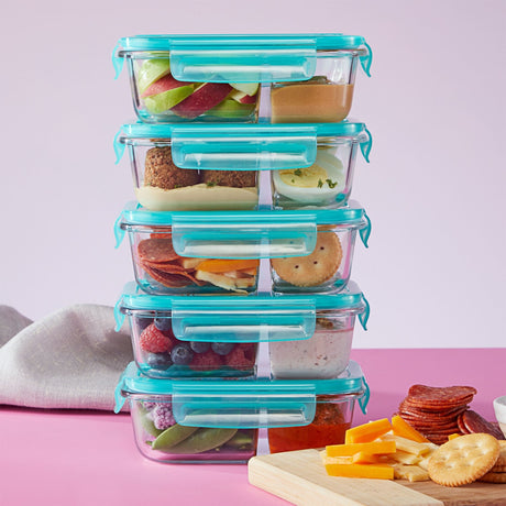 Stack of glass food containers with blue lids filled with various snacks on a pink background