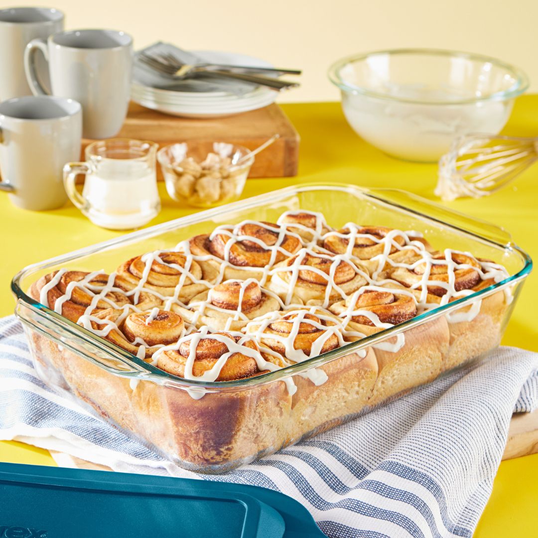 Glass dish with cinnamon rolls on a yellow table with mugs and bowls.
